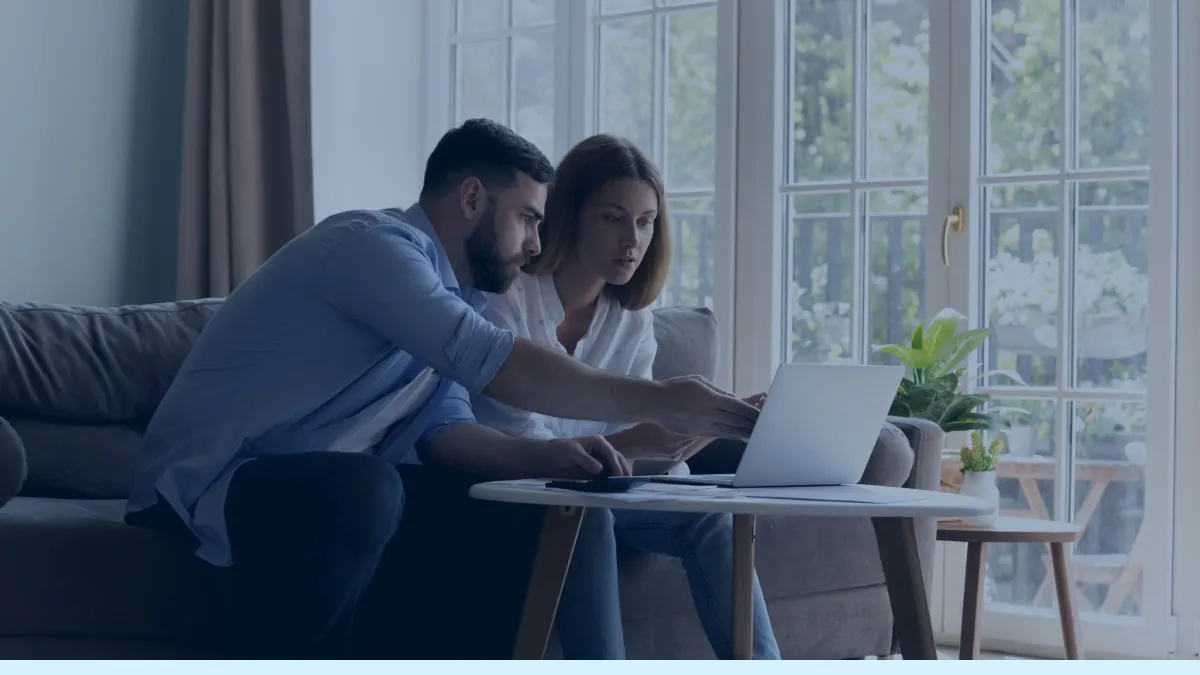 Casal analisando contas em casa, com notebook e calculadora sobre a mesa diante de uma janela